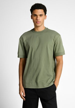Junger Mann mit lockigem Haar, der ein olivgrünes kurzärmeliges T-Shirt und schwarze Hosen trägt, steht mit einer Hand in der Tasche.