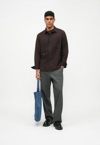 Un uomo in piedi tiene una borsa blu, indossa una camicia button-up marrone scuro, pantaloni grigi e scarpe nere su uno sfondo bianco.