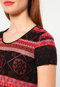 Top en tricot présentant une base noire avec des motifs géométriques multicolores en rouge, rose et argent. Encolure arrondie et manches courtes.