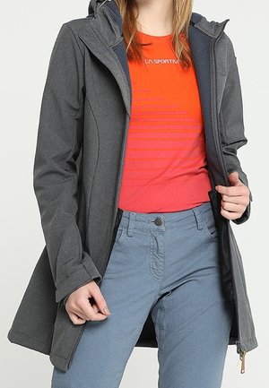 Kvinde iført en mørkegrå hættetrøje over en rød t-shirt med grafik og lyseblå bukser, der holder frakken åben med den ene hånd.