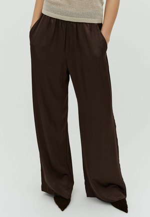 Žena nosi široke hlače tamno smeđe boje s visokim strukom i bočnim džepovima te cipele na špic, uparene s bež džemperom bez rukava.