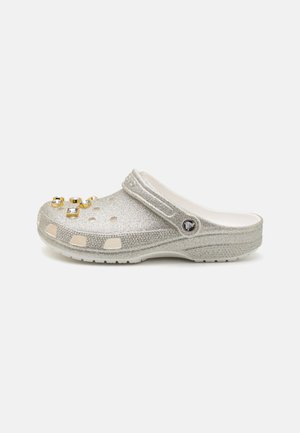 Silver glitter clogs with a rounded toe, ventilation holes, and decorative jewels on the toe. Features a soft inner lining and secure strap.