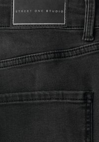 Czarna tkanina denimowa z widocznym szwem, z prostokątną skórzaną metką wytłoczoną z napisem "STREET ONE STUDIO" na obszarze tylnej kieszeni.