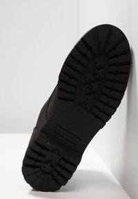 Černá gumová podrážka s hlubokými drážkami, s texturovaným povrchem. Po straně je hladký horní materiál. Vytlačené logo viditelné na podrážce.