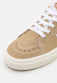 Sneaker in suede beige con chiusura a lacci bianchi, cuciture decorative e dettagli forati sulla punta. Suola in gomma con grip testurizzato.