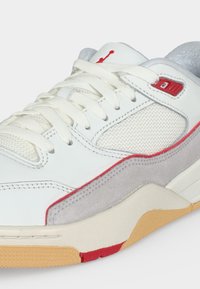 Λευκό και γκρι αθλητικό παπούτσι με διαπνέοντα πάνελ, λευκά κορδόνια, κόκκινες λεπτομέρειες και σόλα χρώματος καουτσούκ, σε παρατήρηση από πλάγια και μπροστινή γωνία.