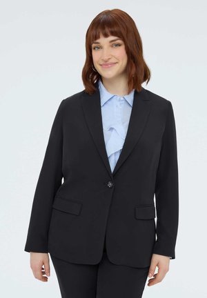 Uśmiechnięta osoba o kasztanowych włosach do ramion, ubrana w czarną marynarkę na jasnoniebieskiej bluzce z falbankami, stojąca na jednolitym tle.