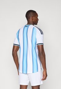 Man die een blauw-wit verticaal gestreept voetbalshirt draagt met het nummer 1893 op de bovenrug en witte korte broek, van achteren gezien.