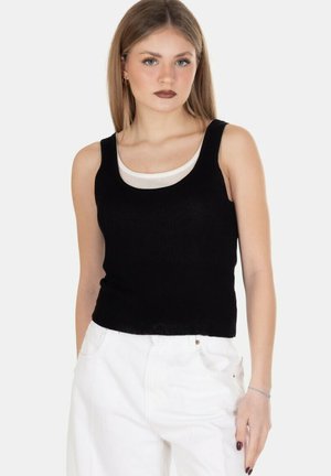 Giovane donna con capelli biondi lisci che indossa un top nero senza maniche sopra una canotta bianca, abbinato a pantaloni bianchi a vita alta, in piedi.