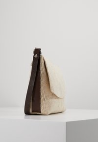 Liten beige vävd tygväska med mörkbrunt läderhandtag och klaff, uppställd på en vit pelare mot en enkel bakgrund.