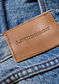 Tessuto di denim con motivo texturizzato azzurro, caratterizzato da un'etichetta in pelle marrone impunturata con la scritta "LINDEBRGH", cucita sulla fascia della vita.