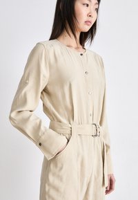 Combinaison en beige à manches longues en tissu doux, avec un col rond, une fermeture à boutons, une ceinture avec boucle et des poches latérales.