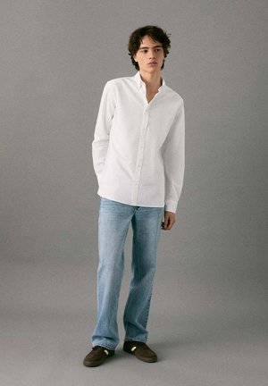 Camicia bianca con bottoni e tessuto morbido, abbinata a jeans azzurri. Il modello è in piedi su uno sfondo grigio neutro, indossando scarpe marroni.