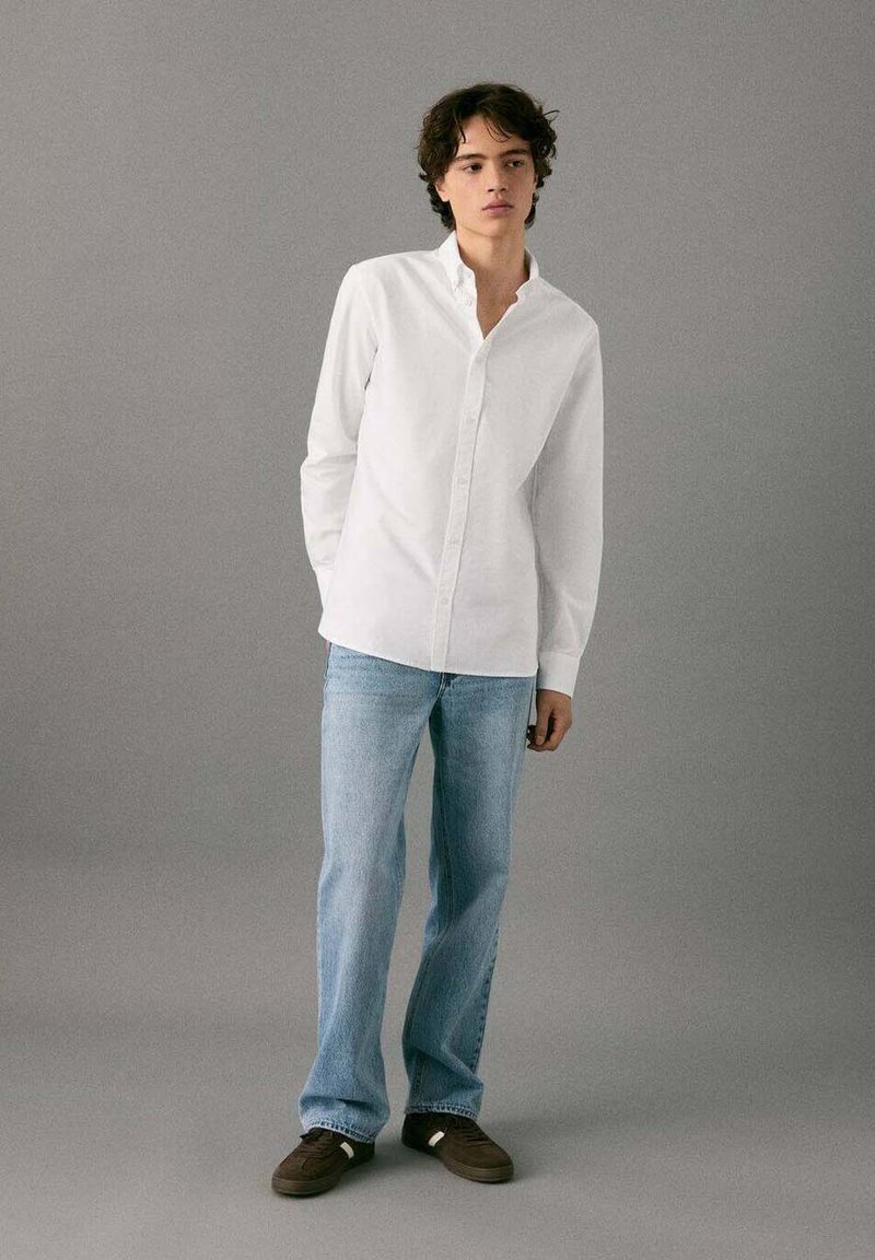 Chemise blanche à boutons avec une texture douce, associée à un jean bleu clair. Le modèle pose devant un fond gris neutre, portant des chaussures marron.