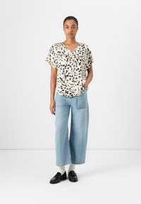Kremfarget bluse med kortermede og svart blomstertrykk, kombinert med lyseblå videjeans og svarte loafers med hvite sokker.