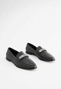 Mustat nahka loaferit, joissa on sileä pinta, kärjelliset muotoilut ja metallinen yksityiskohta ylhäällä. Kapea profiili ja litteä pohja.