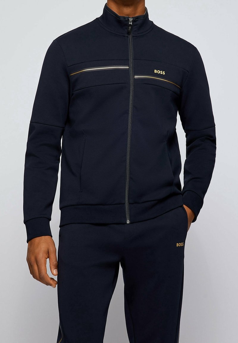 Veste zippée bleu marine en tissu doux. Comprend un col montant, deux accents de bandes horizontales et le logo "BOSS" en or.