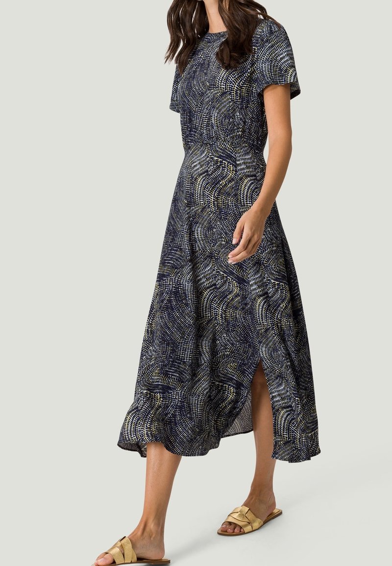 Robe bleu marine avec un motif tourbillonnant en jaune et blanc. Manches courtes, taille cintrée et une fente sur le côté. Portée avec des sandales dorées.