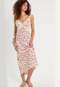 Blumenkleid in Creme mit roten Rosenmustern, Spitzenbesatz am Oberteil, dünnen Trägern und asymmetrischem Saum. Getragen mit goldenen Riemchensandalen.