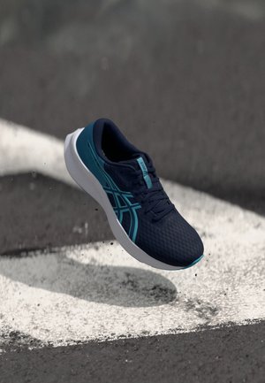 Dunkelblauer Laufschuh mit hellblauen Streifen und weißer Sohle schwebt über Asphalt mit weißen Straßenmarkierungen.