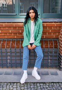 Zelené nadměrné sako, bílé tričko, modré kostkované džíny a bílé tenisky na platformě. Texturované cihlové pozadí a kovové zábradlí.