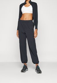 Zwarte sportjack met hoge kraag, witte sport-beha, losse grijze joggers met elastische boorden en zijzakken, en grijze sneakers.