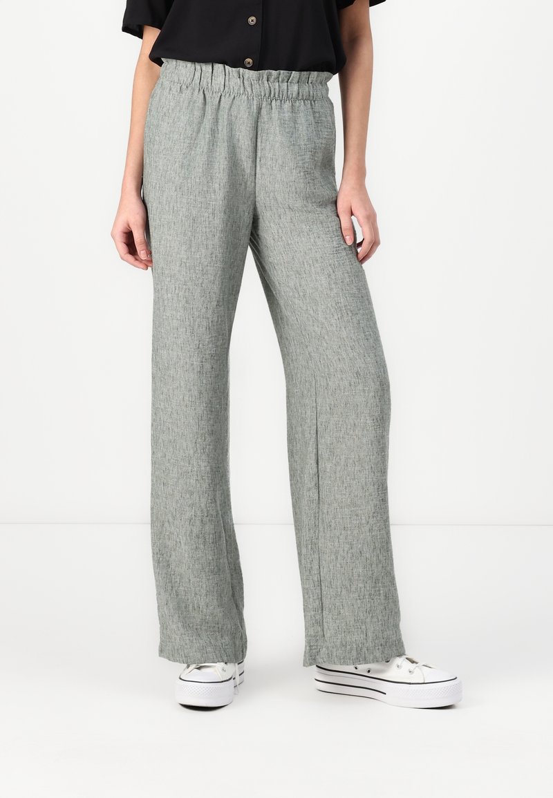 Svetle šedé široké kalhoty s elastickým pasem. Tkanina má texturovaný, tkaný vzhled. Nošeny s bílými teniskami.