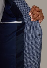 Wnętrze marynarki w granatowo-niebieski wzór pepitki z gładką podszewką, czerwonymi przeszyciami i białą lamówką przy kieszeni.