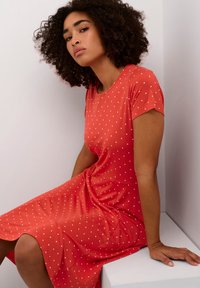 Puna mekko, jossa on valkoisia pilkkuja, lyhyet hihat ja rento istuvuus. Kangas näyttää pehmeältä, siinä on pieniä valumia ja se ulottuu polven ylle.