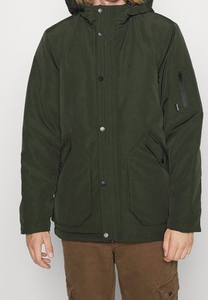 Mężczyzna ubrany w ciemnozieloną zimową kurtkę z kapturem, zapinaną na zatrzaski z przodu, oraz brązowe spodnie, stojący na jednolitym tle.