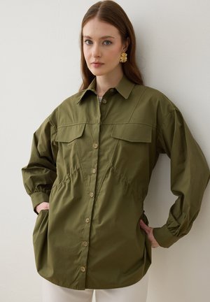 Kvinde med langt brunt hår iført en oversized olivengrøn skjorte med knapper og store guldringe, står mod en hvid væg med hænderne i lommerne.