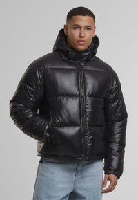 Juodas pūkinis švarkas su blizgiu, siuvinėtu paviršiumi, gobtuvu, užtrauktuku ir šoninėmis kišenėmis, dėvimas ant šviesiai mėlynų džinsų.