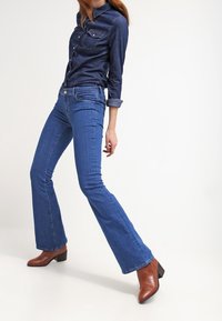 Kvinna klädd i en mörkblå denimskjorta och blå utsvängda jeans med bruna ankelskor, poserar framför en enkel vit bakgrund.