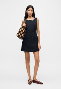 Μπλε σκούρο αμάνικο φόρεμα με κλείσιμο διπλού πέτου με κουμπιά, δύο τσέπες· το μοντέλο κρατά μια καφέ και μπεζ τσάντα με καρό σχέδιο.