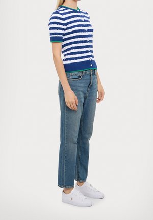 Personne debout en jean bleu décontracté, baskets blanches et cardigan bleu et blanc à rayures, manches courtes, avec liseré vert au col et aux poignets.