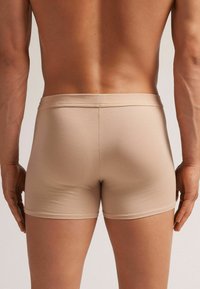 Beige boxer briefs made of soft, stretchable fabric. Features a smooth waistband, fitted design, and minimal seams. Rear view.