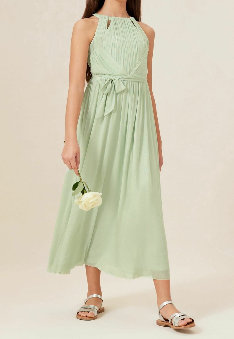 Robe vert clair sans manches avec un corsage plissé, une taille marquée par un nœud et une jupe fluide. Associée à des sandales argentées.