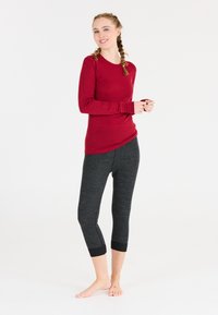 Ung kvinna med flätat hår som bär en röd långärmad topp och mörkgrå croppade leggings, stående barfota mot en vit bakgrund.