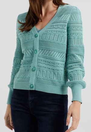 Femme portant un cardigan en maille bleu sarcelle avec des motifs géométriques, boutonné devant et manches longues, sur un jean foncé, sur un fond uni.