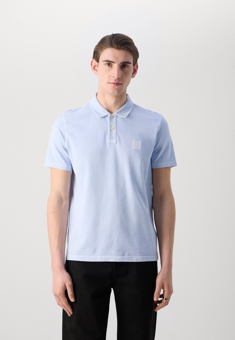 Hellblaues Polohemd aus weichem Baumwollstoff, mit einer Knopfleiste mit drei Knöpfen und einem kleinen Logopatch auf der linken Brust. Kurze Ärmel.