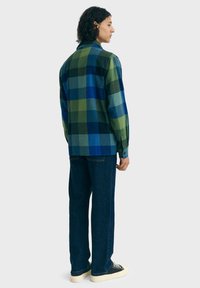 Jongere met krullend haar, gekleed in een blauw en groen geruite shirt, donkere spijkerbroek en zwarte sneakers, staande zijwaarts op een witte achtergrond.