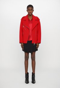 Rote Wollmischung Bikerjacke mit silbernen Reißverschlüssen, über einer roten Bluse getragen. Kombiniert mit einem schwarzen Wickelrock und schwarzen Knöchelstiefeln mit Nieten.