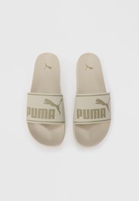 Puma slide sandals in light beige feature a textured band with bold "PUMA" logo and a small jumping cat logo on the footbed. Smooth surface.