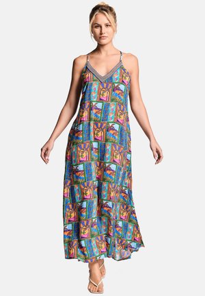 Femme portant une robe maxi patchwork colorée avec des bretelles fines et des motifs tropicaux, debout avec les bras légèrement étendus.