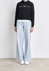 Felpa nera con logo "GANT 1949", camicia a righe azzurre chiaro sotto, jeans larghi azzurri chiari e sneakers bianche. Design semplice e vestibilità rilassata.