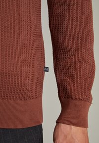 Pull marron tricoté avec un motif texturé en nid d'abeille. Poignets et ourlet côtelés ajoutant un contraste de texture. Petite étiquette visible sur le côté.