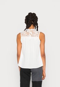 Vit ärmlös topp med en spetsbakpanel, rund halsringning och dekorativ knäppning. Mjukt tyg med en svävande silhuett.