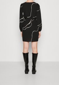 Femme portant une mini-robe noire avec des lignes abstraites blanches, manches longues, et des bottes noires à talons montant jusqu'aux genoux, debout de dos.