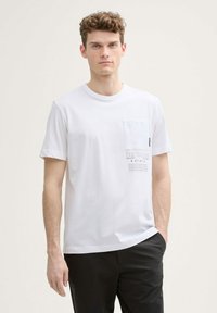 Camiseta blanca de algodón con un bolsillo en el pecho, diseño gráfico impreso en gris y mangas cortas. El modelo lleva pantalones negros.