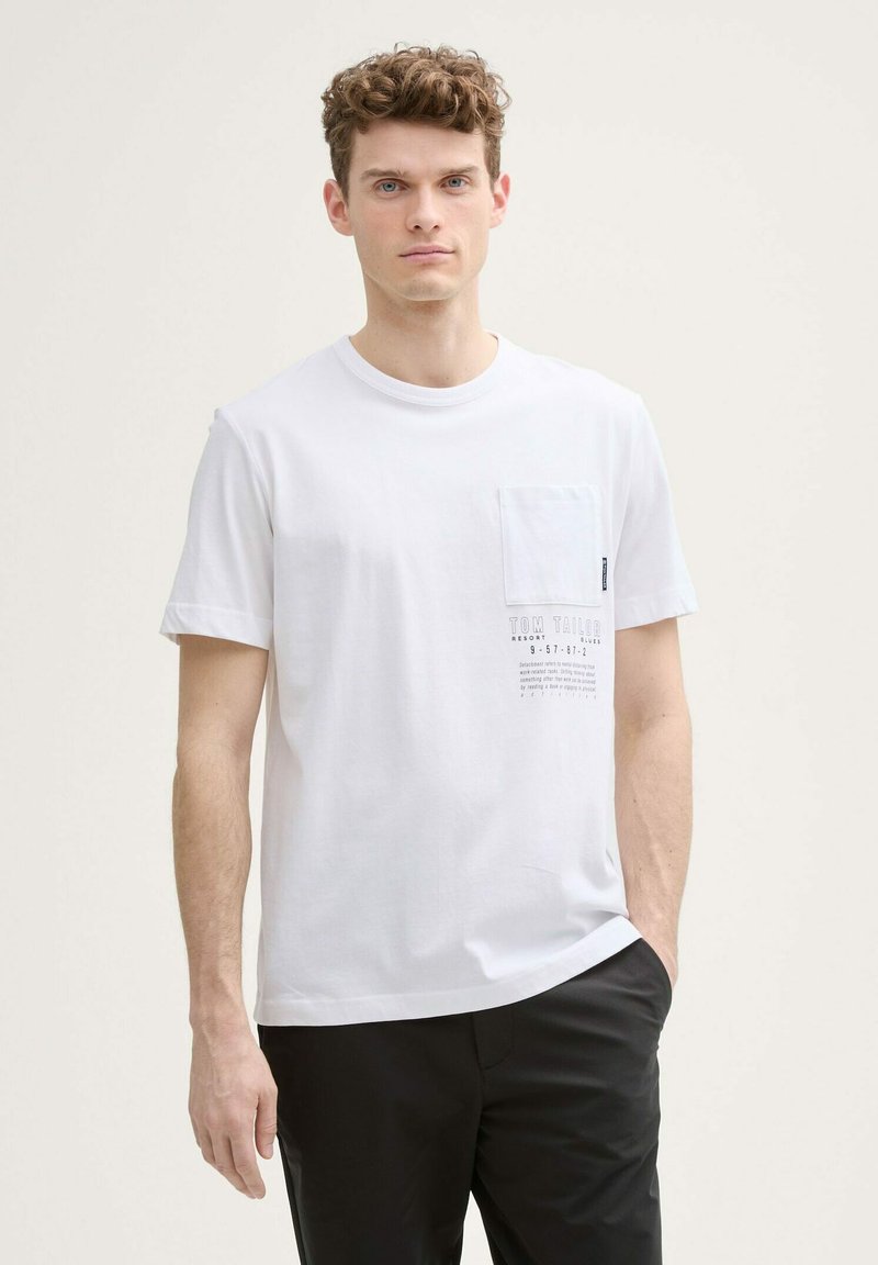 Camiseta blanca de algodón con un bolsillo en el pecho, diseño gráfico impreso en gris y mangas cortas. El modelo lleva pantalones negros.